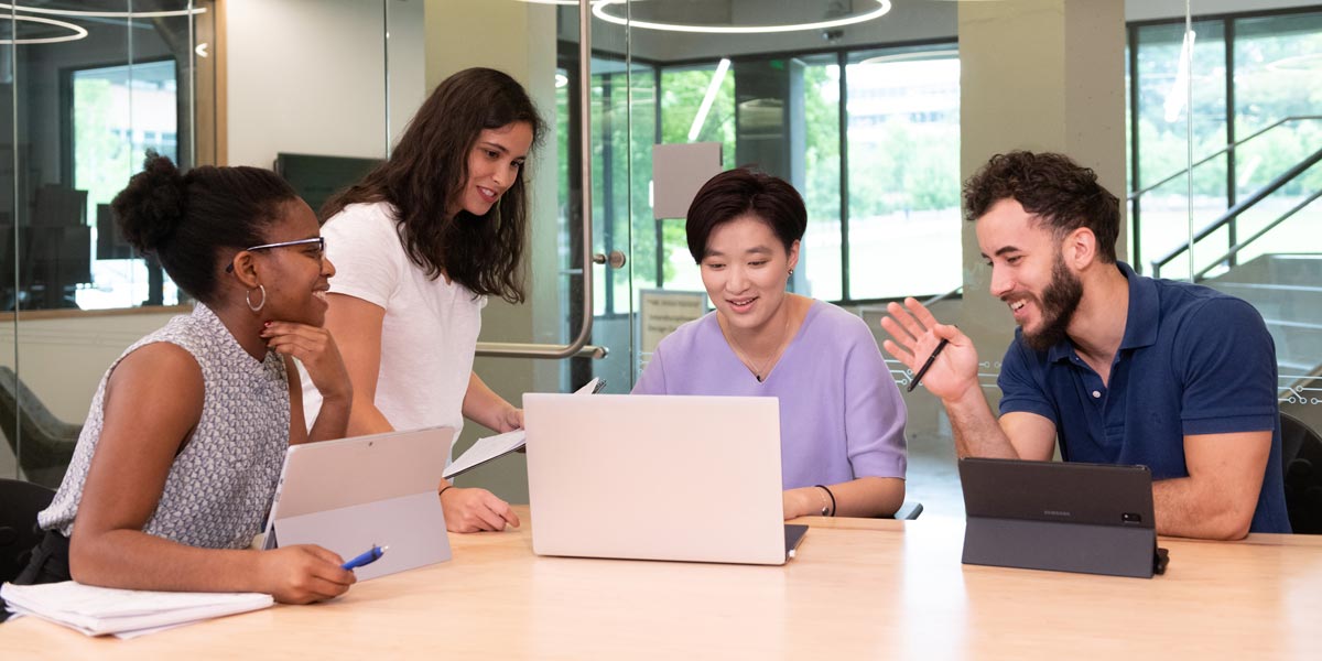 Four students sitting at a table working on laptops and tablets.