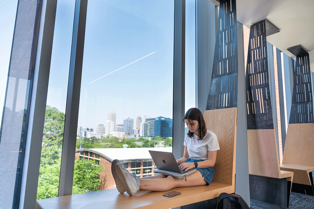 A student sits on a bench next to a window with a laptop on her lap